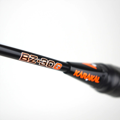 Close-up of the Karakal Black Zone BZ 30 2.1 badminton racket handle in black with orange lettering, "KARAKAL," on a blurred white background, highlighting its isometric head design.