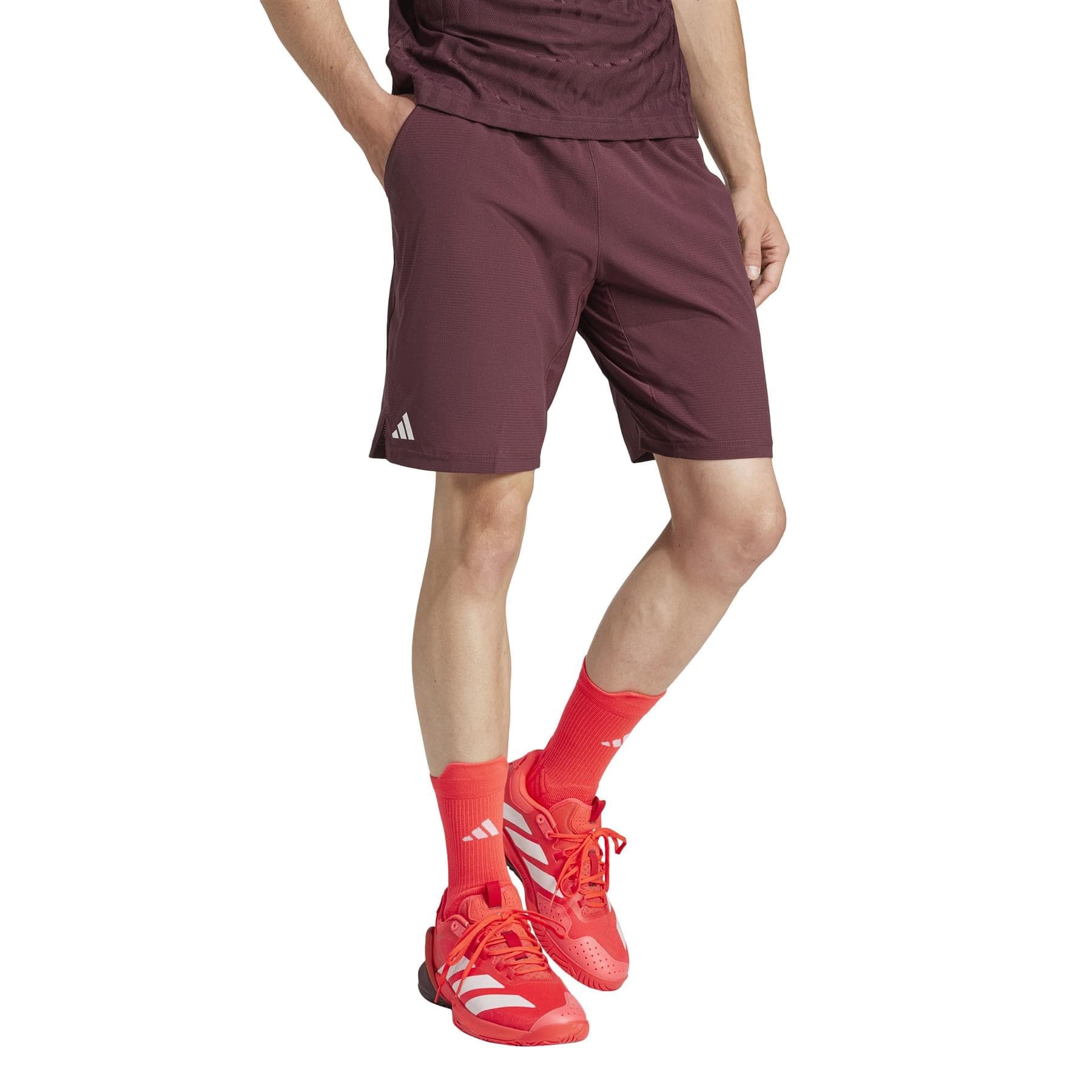 Someone is shown wearing ruby ADIDAS Ergo Men's Badminton Shorts by adidas, made from recycled materials, with a matching top and red athletic shoes featuring white accents. Red socks complete the sporty look, while the photo emphasizes their lower body attire.