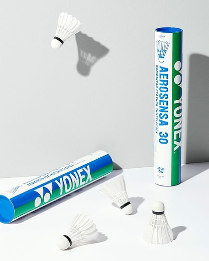 Yonex Aerosensa AS30 Feather Shuttles (Set of 12) by Yonex are shown on a light surface—two shuttlecocks appear mid-air, highlighting flight stability, with two branded tubes displayed, one upright and one lying down.