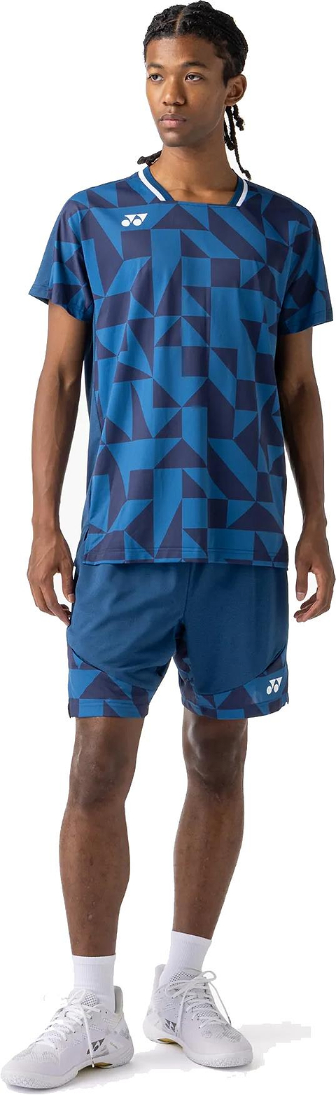 A man wearing a Yonex 10712YX Crew Neck Men's Badminton T-Shirt in Indigo Blue with matching shorts, white socks, and sneakers stands slightly turned to the side against a white background.