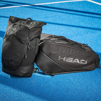 Two black HEAD Pro X Badminton Backpack 25L bags rest on a blue tennis court. Their modern urban style and prominent HEAD logo stand out sharply against the white lines in the background.