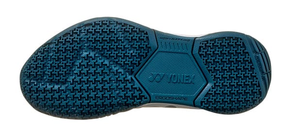 The image shows the sole of a Yonex Power Cushion 65 Junior Badminton Shoe, featuring a textured blue rubber with hexagonal and cross shapes. It includes "NON-MARKING," "ERGO-SHAPE," and the Yonex logo, boasting Power Cushion technology for superior shock absorption.