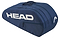 A blue HEAD BASE 6 Racket Badminton Bag M - NV, featuring the brand in large white letters, has a curved design with multiple compartments and zippers, reinforced for durability, a top handle, and holds up to six rackets.