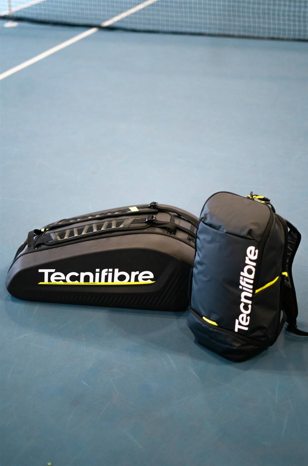 A large black Tecnifibre racket bag and a Tecnifibre Reform Badminton Backpack - Black are placed on a blue tennis court near the net.