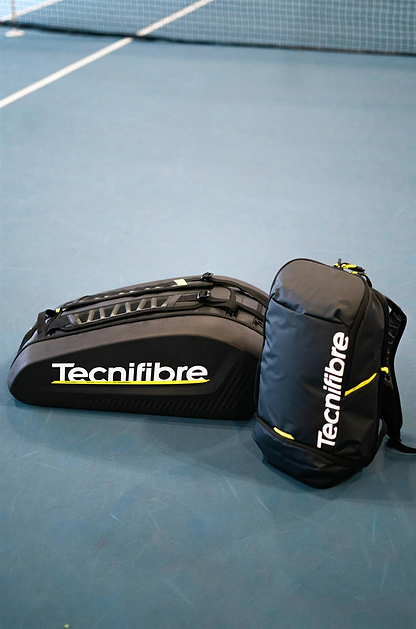 A large black Tecnifibre racket bag and a Tecnifibre Reform Badminton Backpack - Black are placed on a blue tennis court near the net.