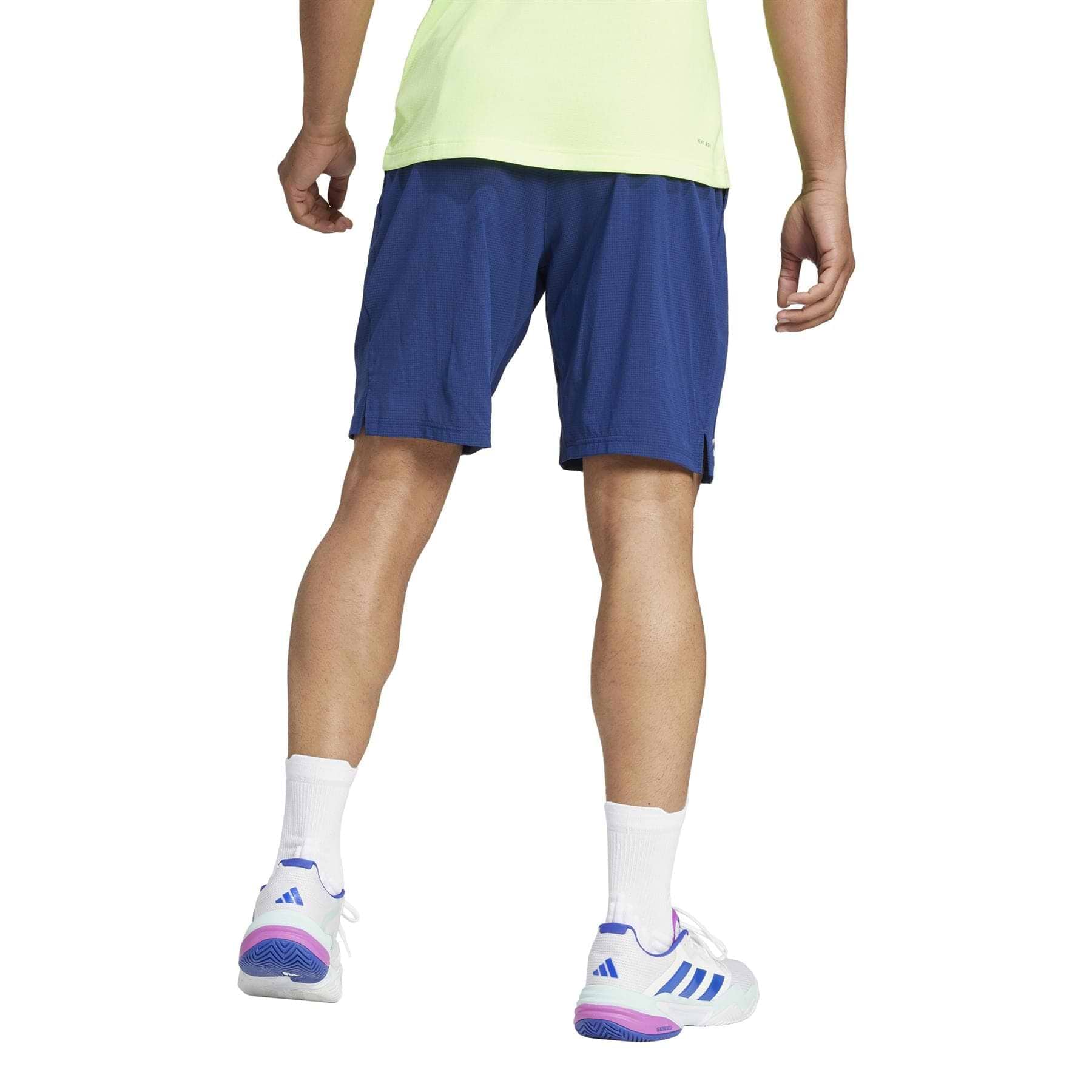 A person in a light green shirt, navy ADIDAS Ergo Mens Badminton Shorts made of recycled materials, and white sneakers with purple and blue accents stands facing away, their white socks complementing the athletic footwear against a plain white backdrop.