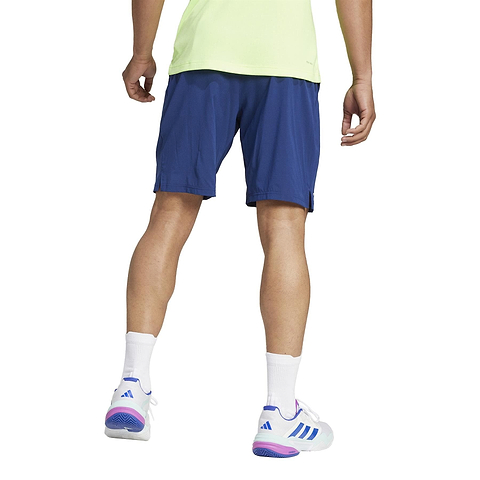 A person in a light green shirt, navy ADIDAS Ergo Mens Badminton Shorts made of recycled materials, and white sneakers with purple and blue accents stands facing away, their white socks complementing the athletic footwear against a plain white backdrop.