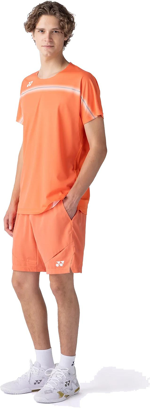 A young man stands with one hand in his pocket, wearing the Yonex 10711YX Crew Neck Mens Badminton T-Shirt in Cyber Orange—premium Yonex performance apparel—paired with matching shorts and white athletic shoes against a plain white background.
