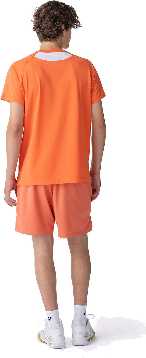 A person with curly hair stands facing away, wearing the Yonex 10711YX Crew Neck Mens Badminton T-Shirt in Cyber Orange, matching shorts, white socks, and white sneakers against a white background.