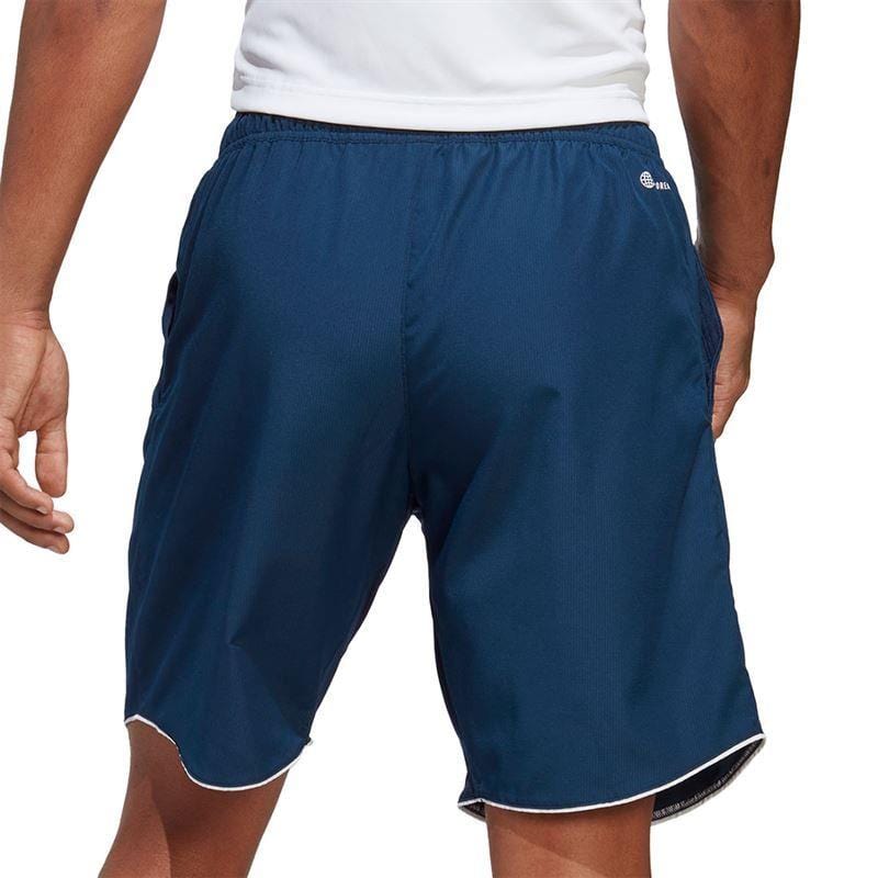The image shows a person from the waist down, wearing a white shirt paired with navy ADIDAS Mens Club 7" Badminton Shorts. Made by adidas, these shorts are equipped with AEROREADY fabric for effective moisture wicking. They feature a small logo on the back and a white trim along the edges, as the individual stands outdoors.