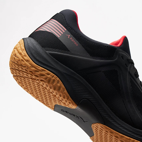 Close-up of the black Kanso Equilibrium EQx3 Unisex Badminton Shoe, featuring orange-brown soles with Speedlight foam for comfort, red heel accents, and a textured tread. The shoe is angled with the sole visible against a white background.