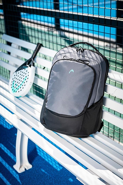 A HEAD Pro X Badminton Backpack 30L in Black/Dark Grey rests on a white bench beside a padel racket with a white face and black handle. The bench is near a black wire fence on the blue court, and the eco-friendly setup, made from recycled PET bottles, casts shadows in bright daylight.
