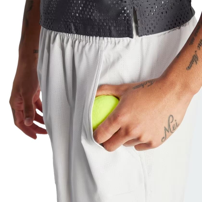 A person is holding a tennis ball in their right hand, with it partially tucked into the side pocket of adidas Melbourne Shorts & Inner Shorts Men's Badminton Set in Grey One/Carbon. They are wearing a dark, perforated top featuring HEAT.RDY technology and have tattoos on their left wrist and hand.