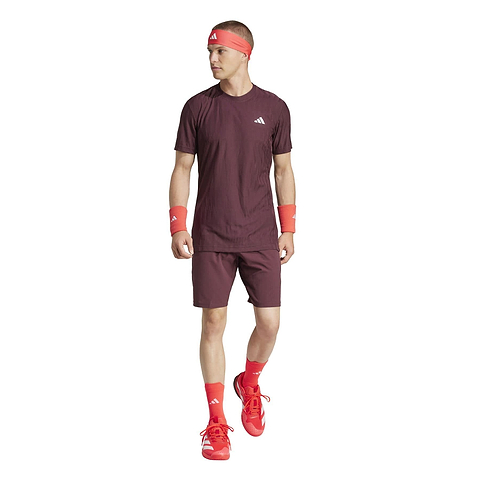 On a white background, a person confidently wears adidas Ergo Men's Badminton Shorts in Ruby, complemented by a maroon athletic set including a matching T-shirt, red sneakers, headband, wristbands, and socks while gazing slightly to the side.