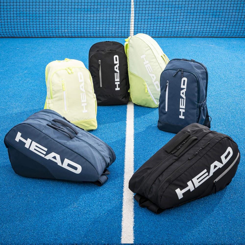 Six HEAD BASE Badminton Backpack 17L - Black bags are arranged in a semicircle on a blue tennis court near the net—three upright and three lying down.