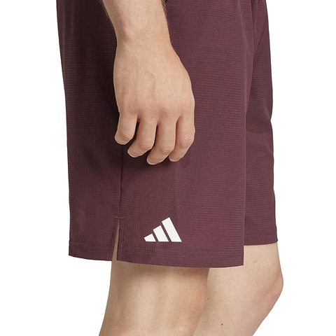 Someone is wearing ADIDAS Ergo Men's Badminton Shorts in Ruby, displaying part of their hand and leg. These maroon shorts have a white adidas logo, a small side slit for mobility, and feature HEAT.RDY technology to keep you cool during intense matches.