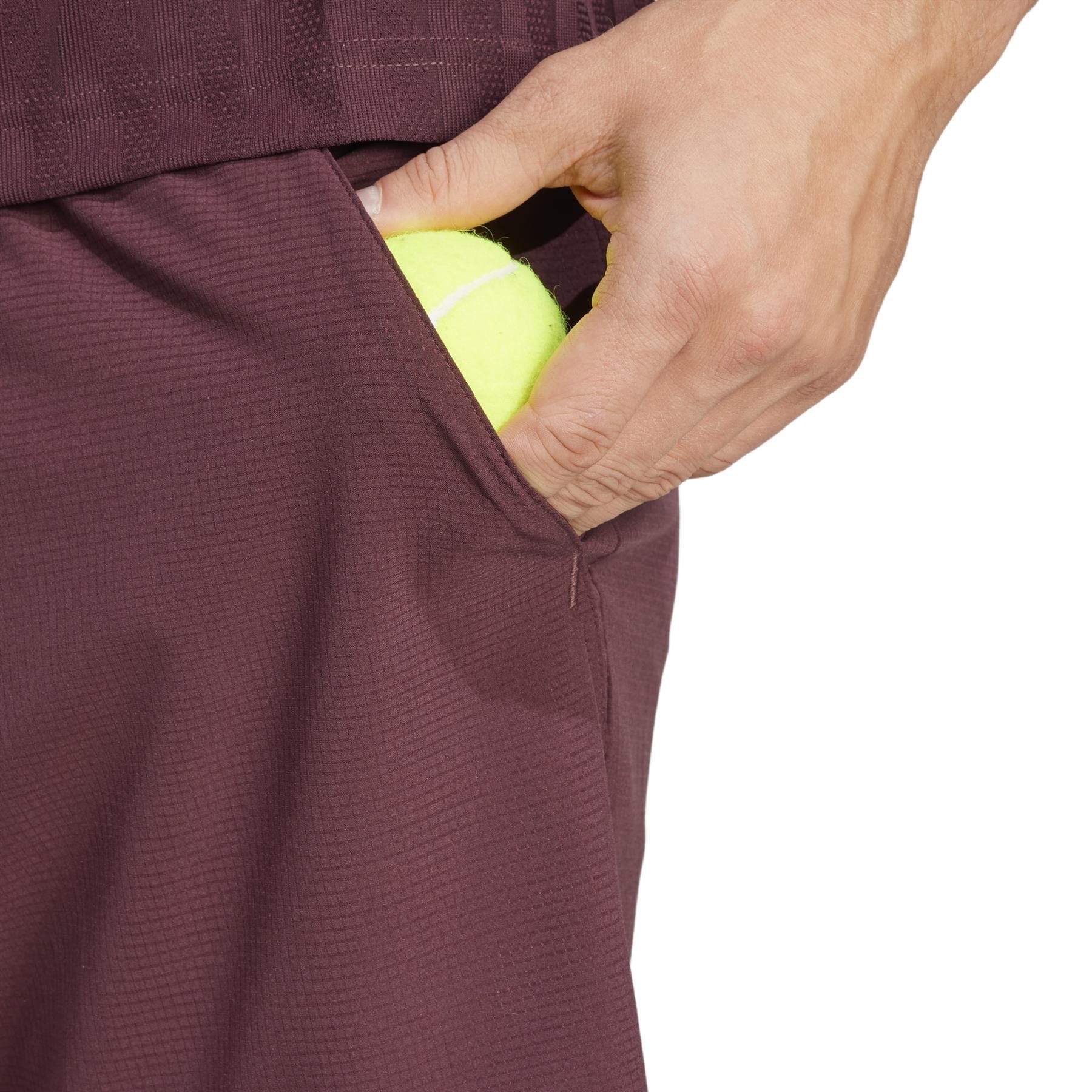 A person wearing adidas ADIDAS Ergo Men's Badminton Shorts in Ruby, made from recycled materials, holds a yellow tennis ball partially tucked into the pocket. The focus is on their hand and the tennis ball.