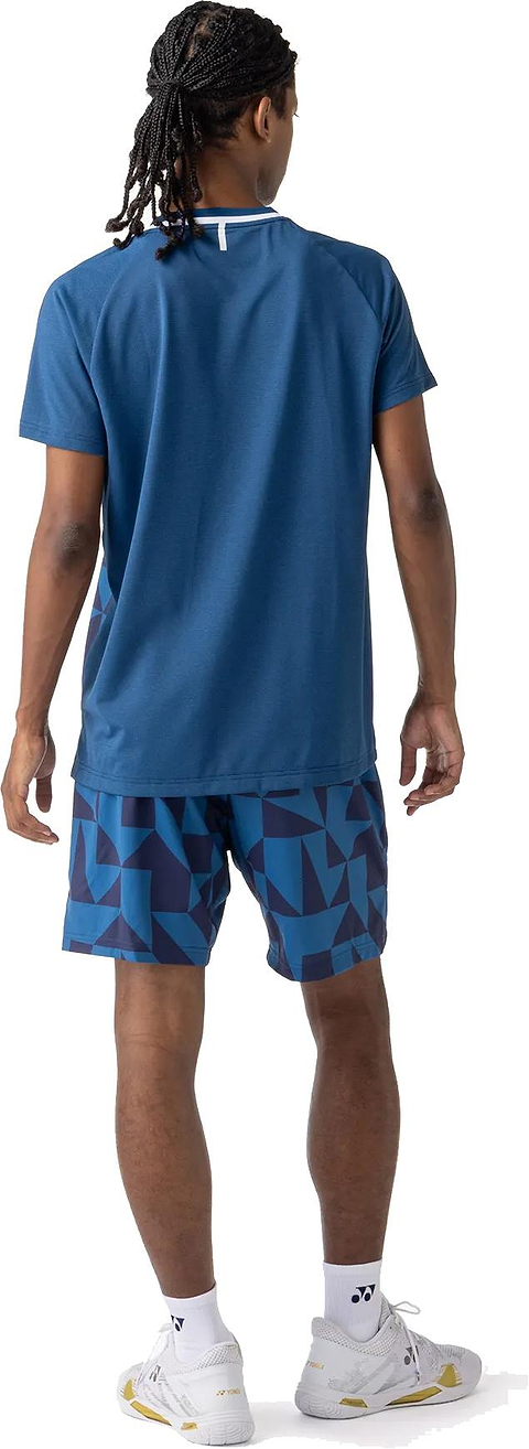 A person with braided hair is standing and facing away, wearing a blue T-shirt, Yonex 15240YX Knit Mens Badminton Shorts in Indigo Blue, white socks, and white athletic shoes.