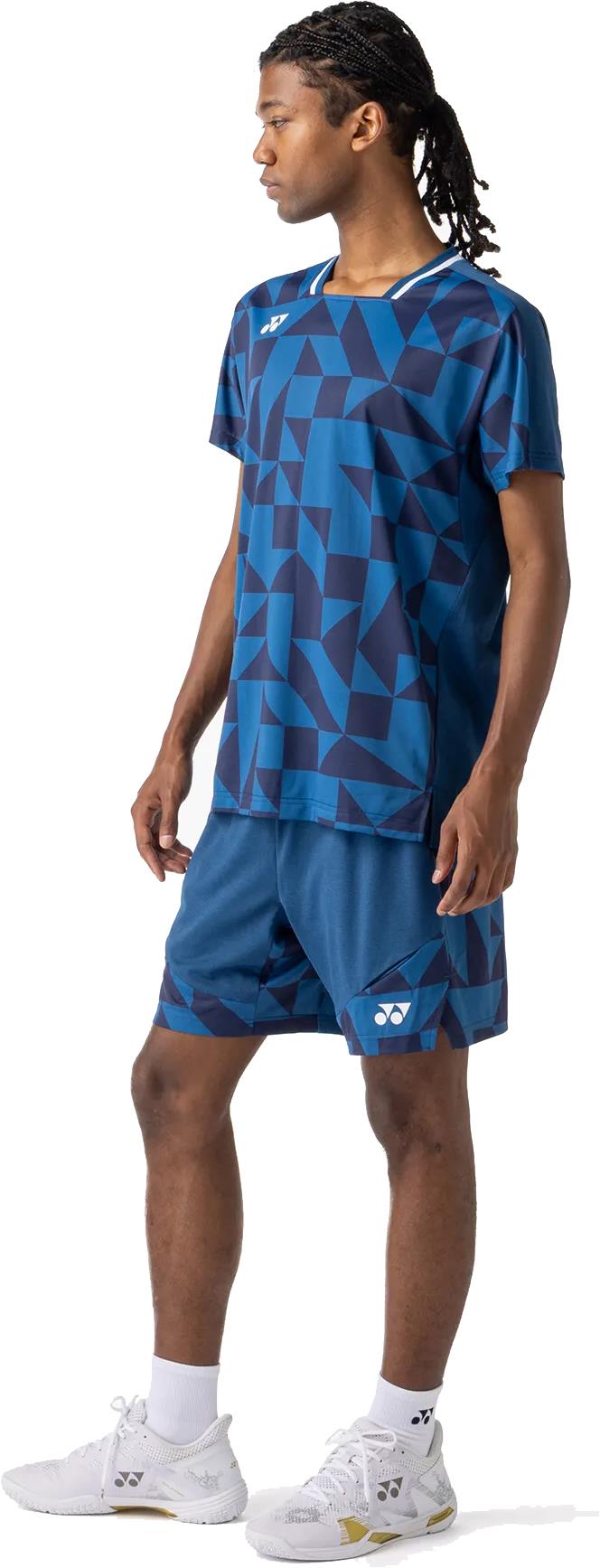 A person wearing a Yonex 10712YX Crew Neck Men's Badminton T-Shirt in indigo blue stands facing left, paired with matching shorts, medium-length braided hair, white socks, and white athletic shoes.