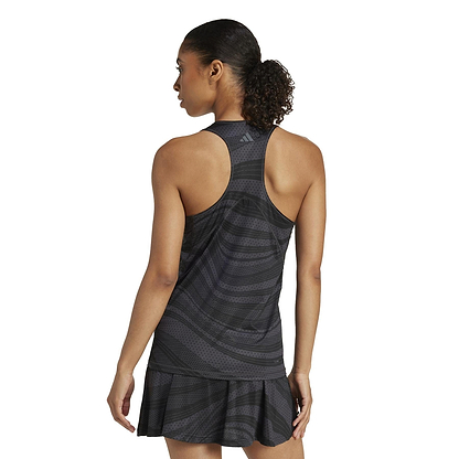 A curly-haired person in a sporty, sleeveless black dress stands facing away, looking to the side. The dress, made from recycled materials, has a racerback style and pleated skirt similar to the ADIDAS Women's Club Graphic Badminton Tank Top. The background is white.