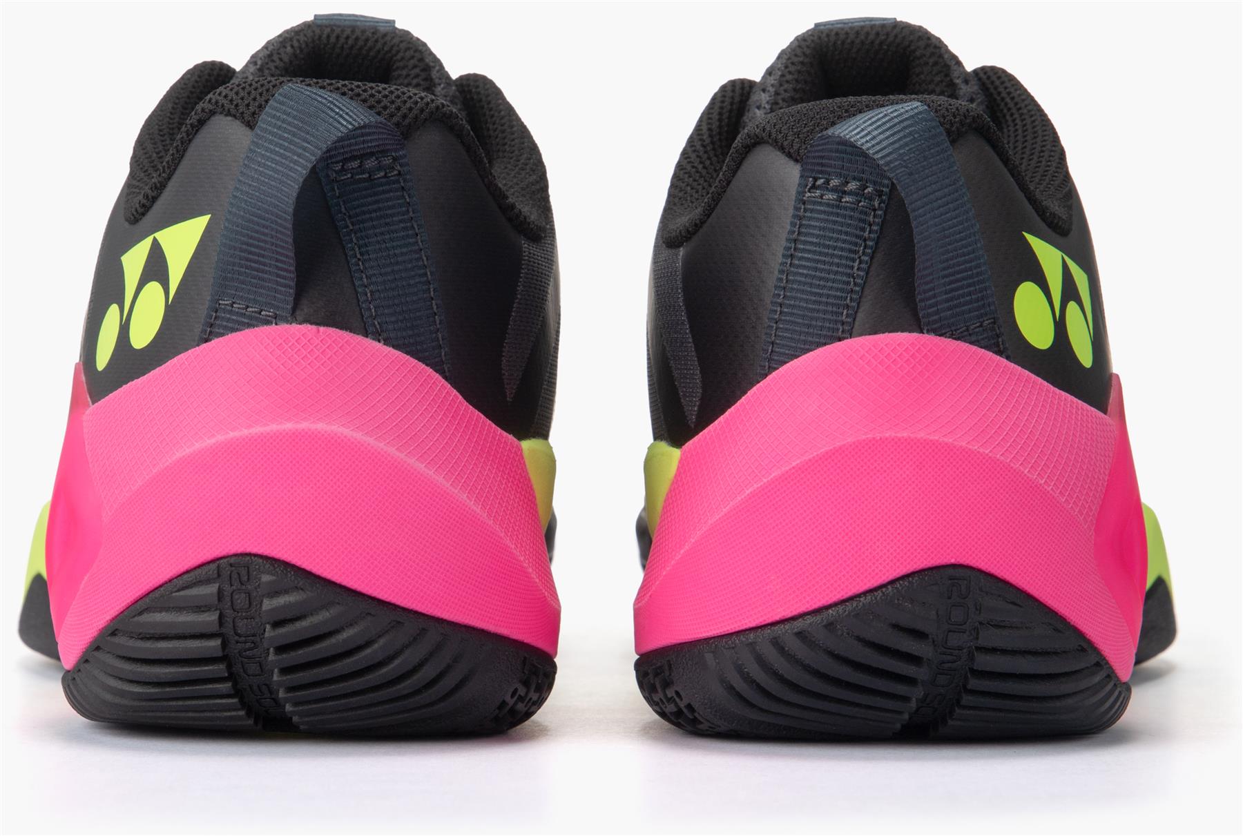 A rear view of two Yonex Subaxia GT Mens Badminton Shoes in dark grey, with neon yellow logos, bright pink heel accents, black GRPHT THRTTL soles, and dark blue pull tabs, displayed on a white background.