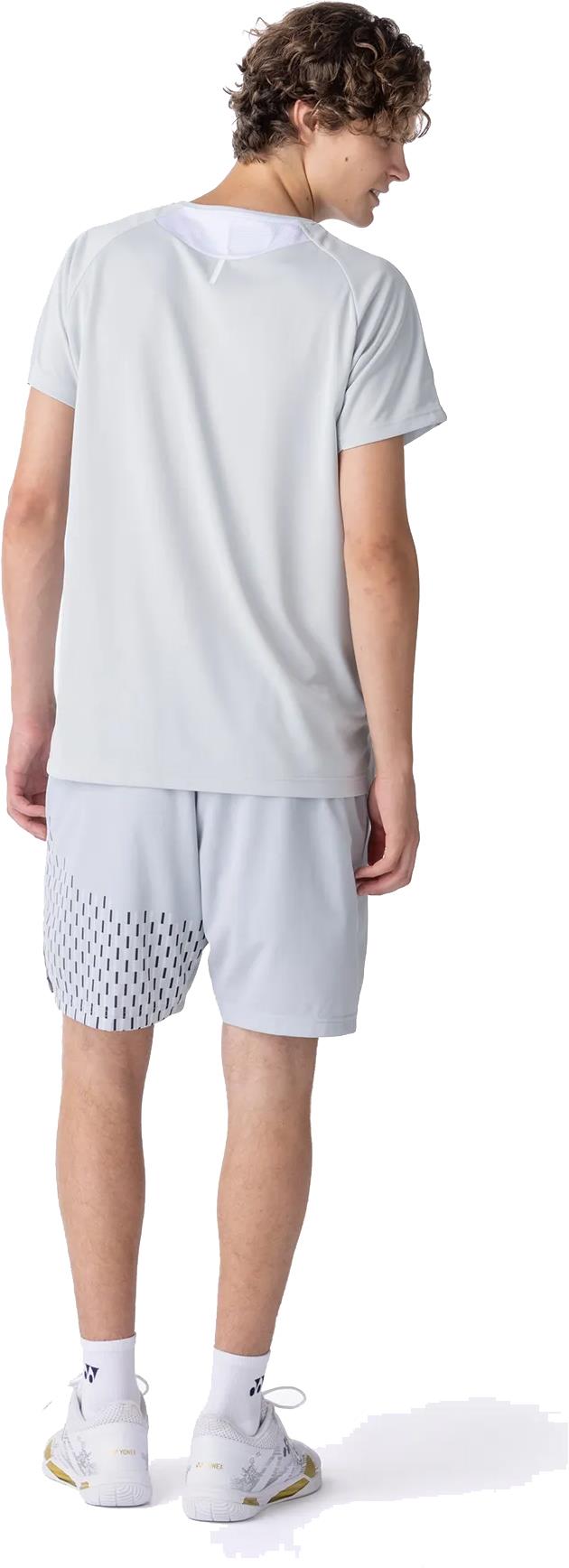 A person with curly hair wears a Yonex 10713EX Crew Neck Men's Badminton T-Shirt in Silver Grey and patterned gray athletic shorts, white socks, and white sneakers, standing with their back to the camera against a white background.
