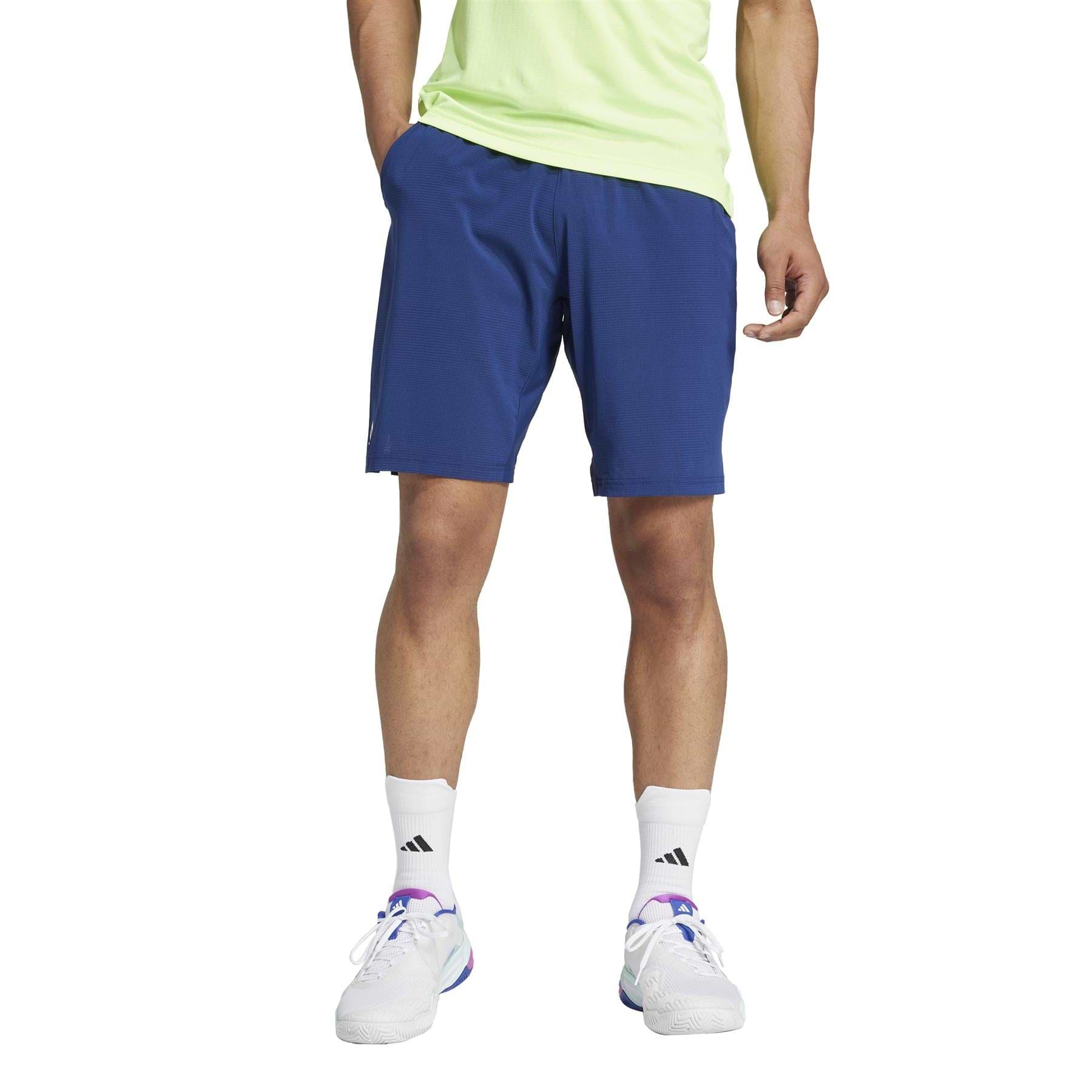 A person wearing a light green shirt and ADIDAS Ergo Men's Badminton Shorts in navy stands against a plain background. They sport white HEAT.RDY socks and sneakers with purple accents, with their hands resting casually in their pockets.