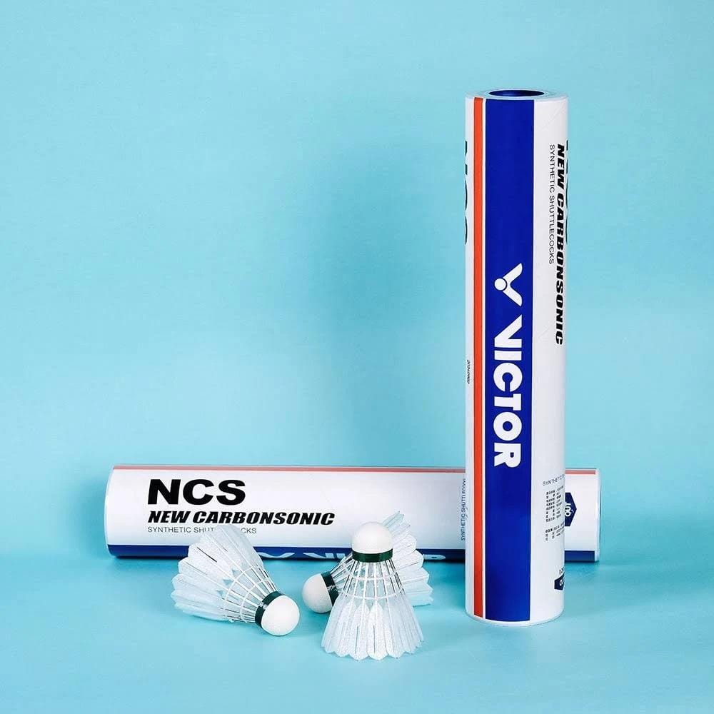 Three white synthetic feather Victor New Carbonsonic NCS Badminton Shuttlecocks are placed in front of two Victor-branded tubes, one upright and one lying down, all set against a light blue background. Set contains 12 shuttlecocks.