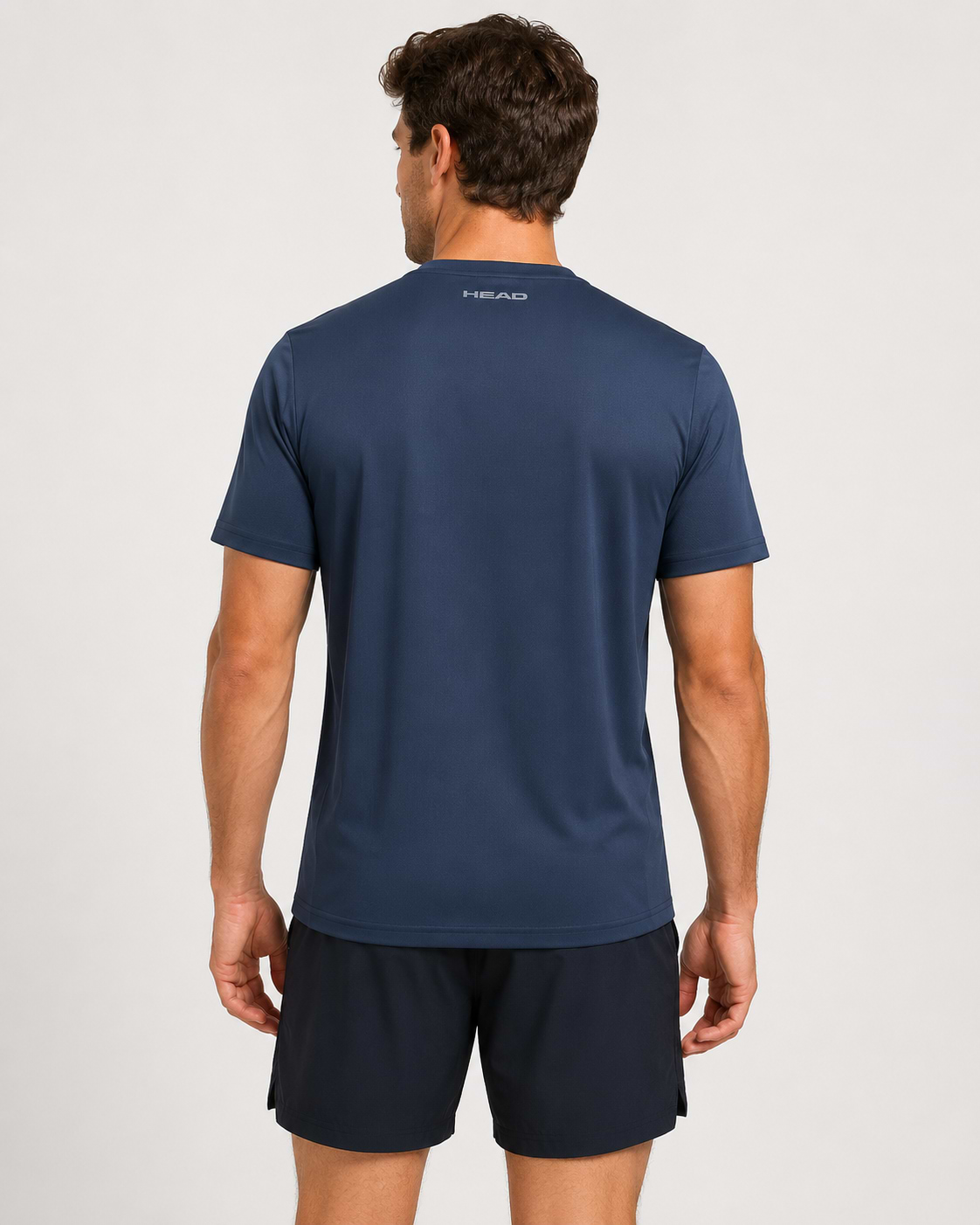 A man, facing away from the camera in black shorts, wears a navy HEAD Club Tech 25 Men’s Badminton T-Shirt by HEAD. This breathable shirt with moisture transfer microfiber has "HEAD" printed on the upper back against a light background.