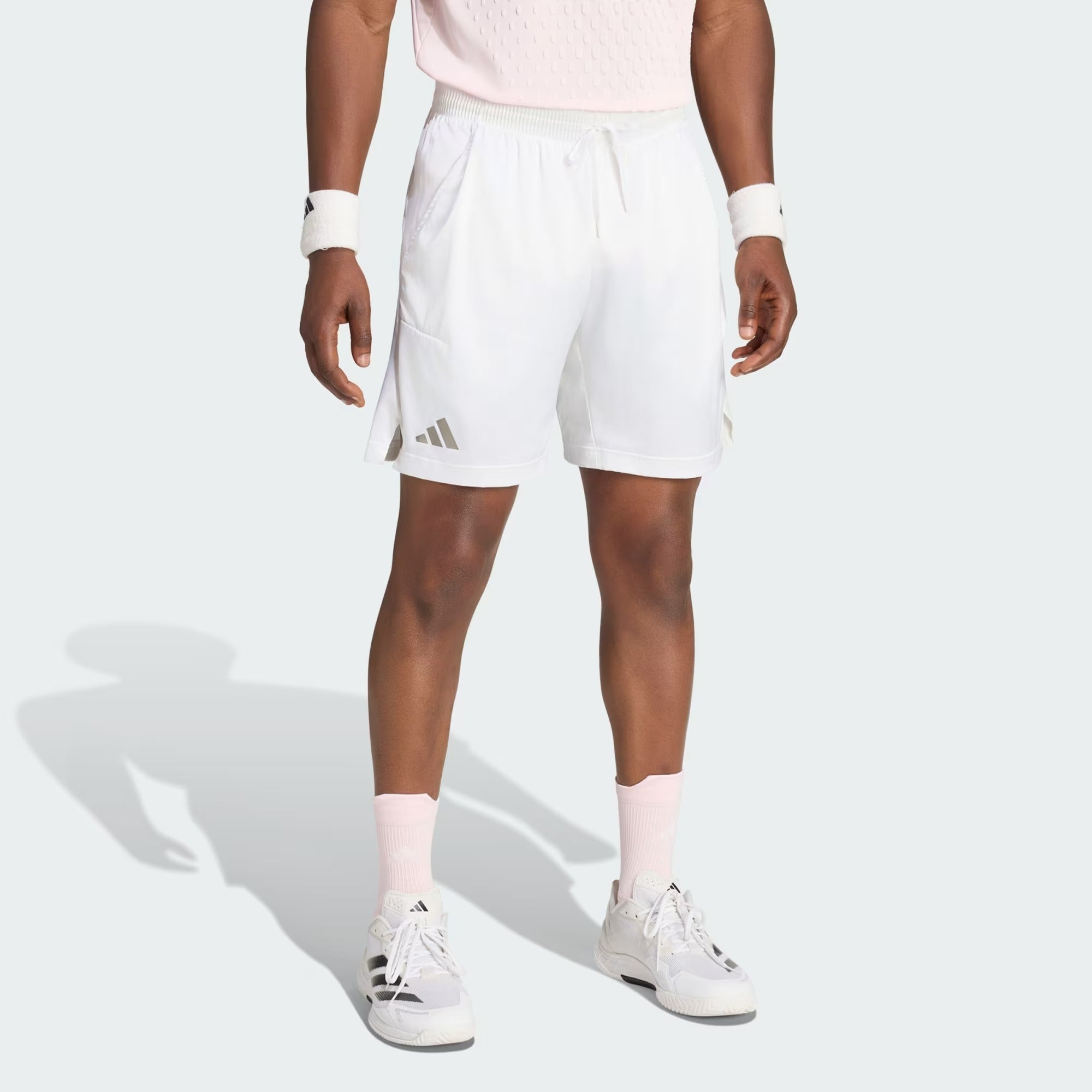 A person wearing adidas ADIDAS Climacool Ergo Pro Men’s Badminton Shorts in white, along with white and pink sneakers, pink socks, a pink shirt, and white wristbands, is standing against a light background. Only the lower half of their body is visible.