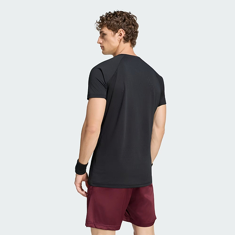 A man with curly hair, dressed in a black adidas Climacool+ Freelift Pro Men’s Badminton T-Shirt—engineered with advanced cooling technology—along with maroon shorts and a black wristband, stands facing away from the camera against a plain light background.