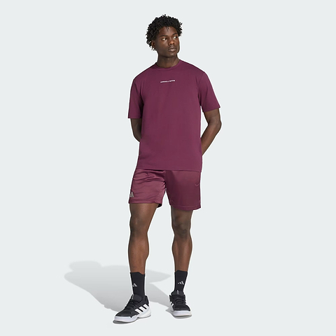 A man stands against a light background wearing a matching maroon adidas t-shirt and ADIDAS Climacool Ergo Pro Men’s Badminton Shorts in maroon. He pairs these with black socks and black-and-white adidas sneakers, has his hands behind his back, and looks slightly to the side.