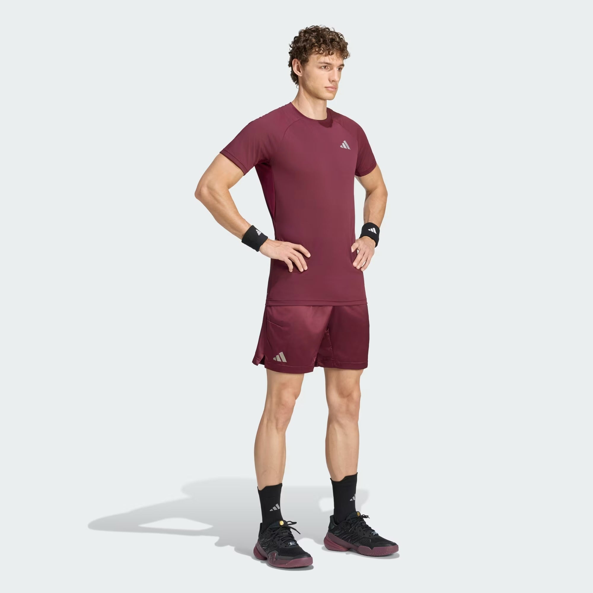 A man stands with hands on hips, wearing the adidas Climacool+ Freelift Pro Men’s Badminton T-Shirt in maroon, along with matching shorts, black wristbands, socks, and shoes—all featuring adidas logos—set against a light gray background.