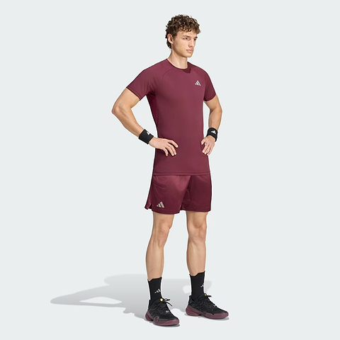 A man stands with hands on hips, wearing the adidas Climacool+ Freelift Pro Men’s Badminton T-Shirt in maroon, along with matching shorts, black wristbands, socks, and shoes—all featuring adidas logos—set against a light gray background.