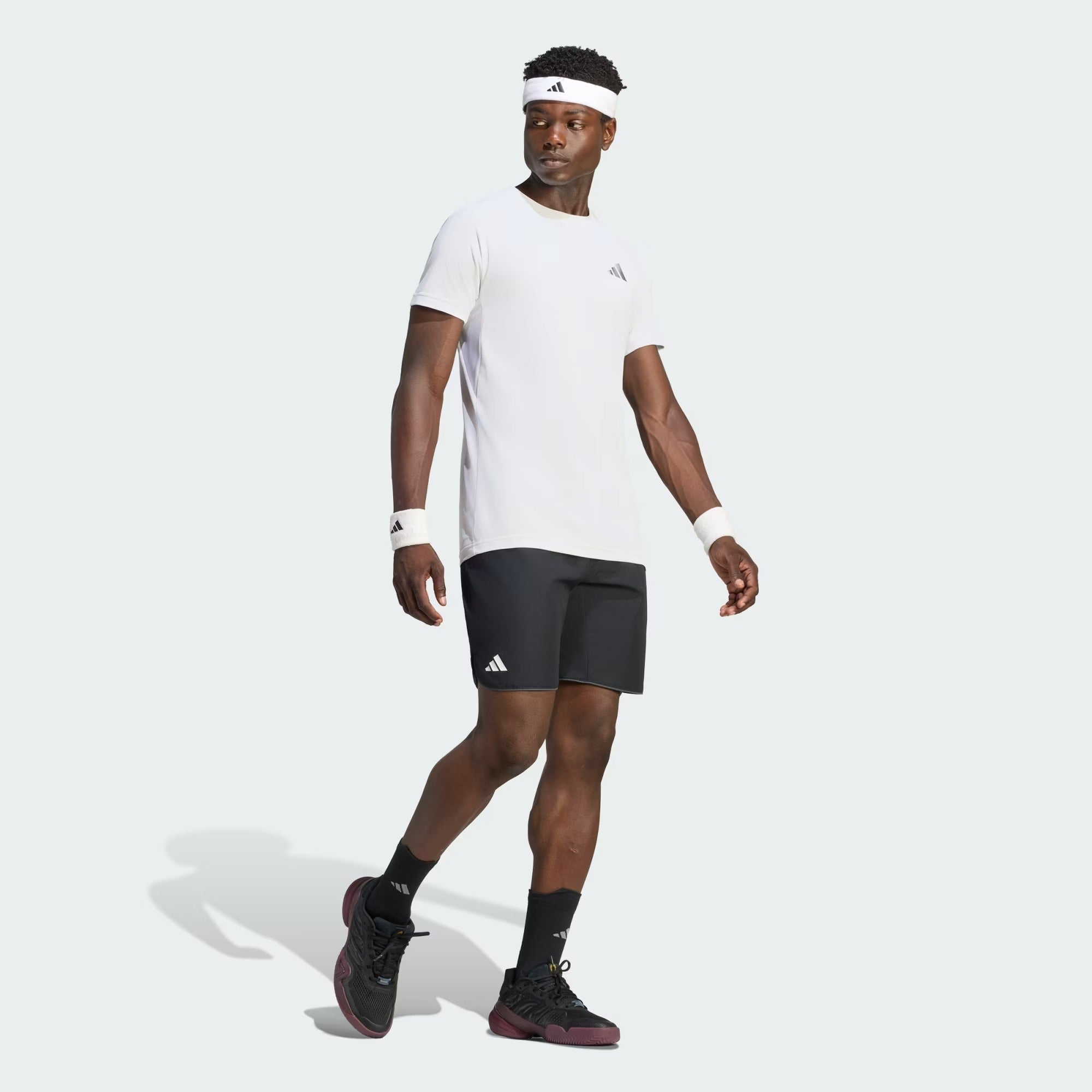 A man stands confidently against a plain light gray background, dressed in the ADIDAS Climacool+ Freelift Pro Men’s Badminton T-Shirt in white by adidas, paired with black shorts, black socks, and black sneakers. He accessorizes with a white headband and wristbands. The quick-drying sports tee ensures comfort during intense activity.