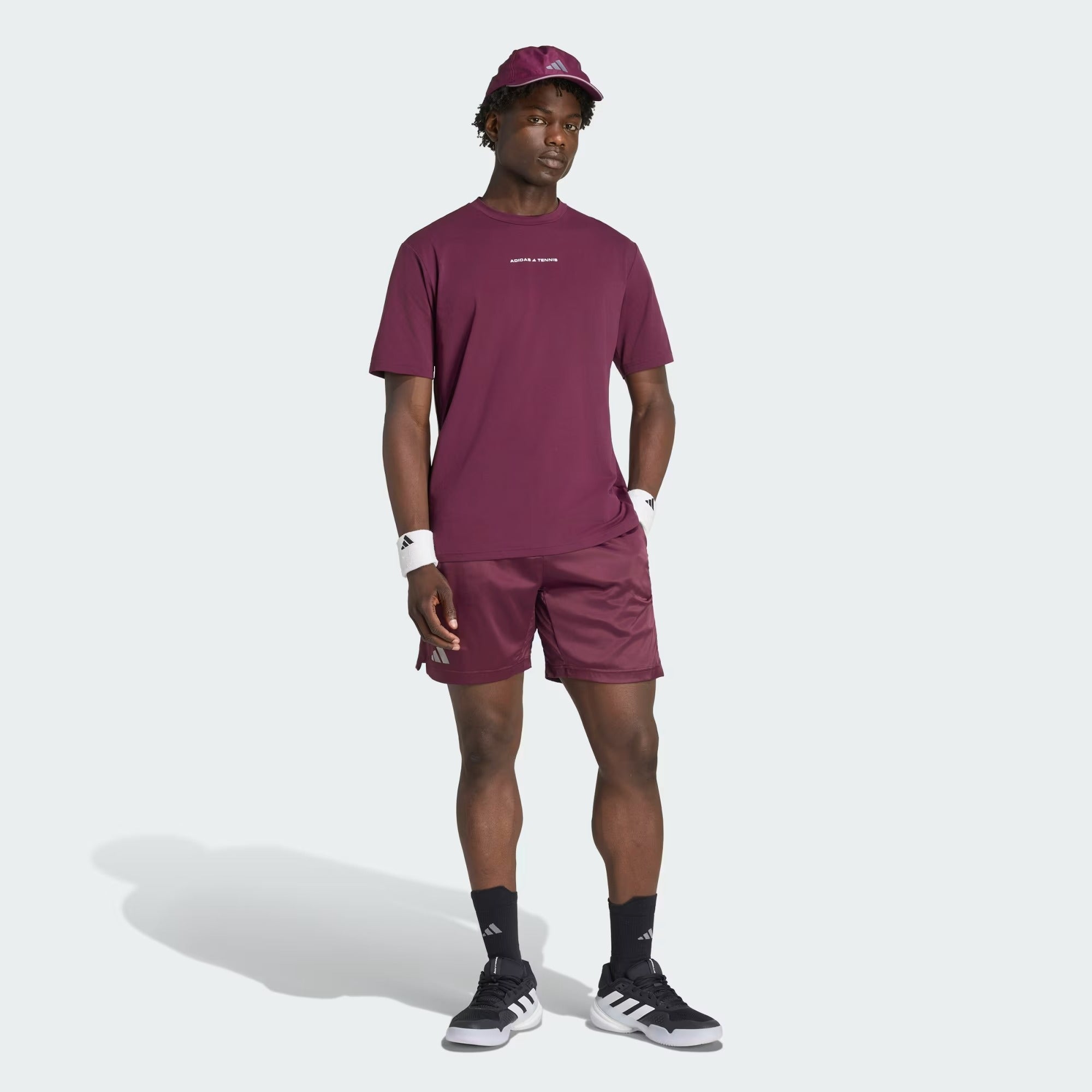 A man wearing a maroon t-shirt, matching adidas Climacool Ergo Pro Men’s Badminton Shorts in maroon, a cap, white wristbands, black socks, and black adidas sneakers stands with one hand in his pocket against a plain white background. The ergonomic design of the Climacool Ergo Pro shorts provides optimal comfort and style for any active look.