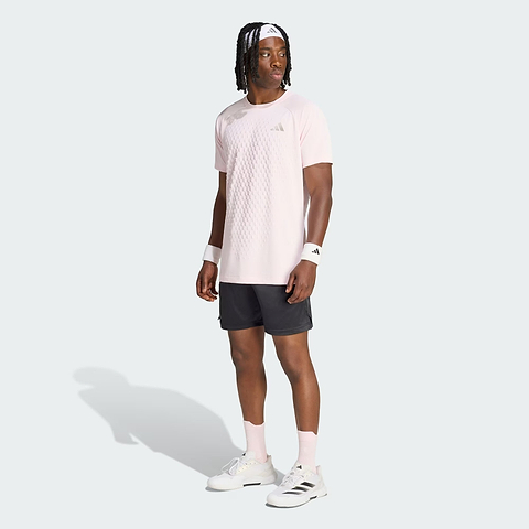 A man wearing a light pink Adidas shirt, black adidas Climacool Ergo Pro men’s badminton shorts, white wristbands, white socks, and white adidas sneakers stands facing slightly left against a plain light background.