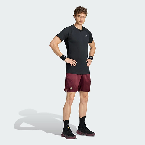 A person stands with hands on hips, wearing an adidas Climacool+ Freelift Pro Men’s Badminton T-Shirt in black, featuring advanced cooling technology, maroon shorts, black shoes, black wristbands, and black socks against a plain light background.