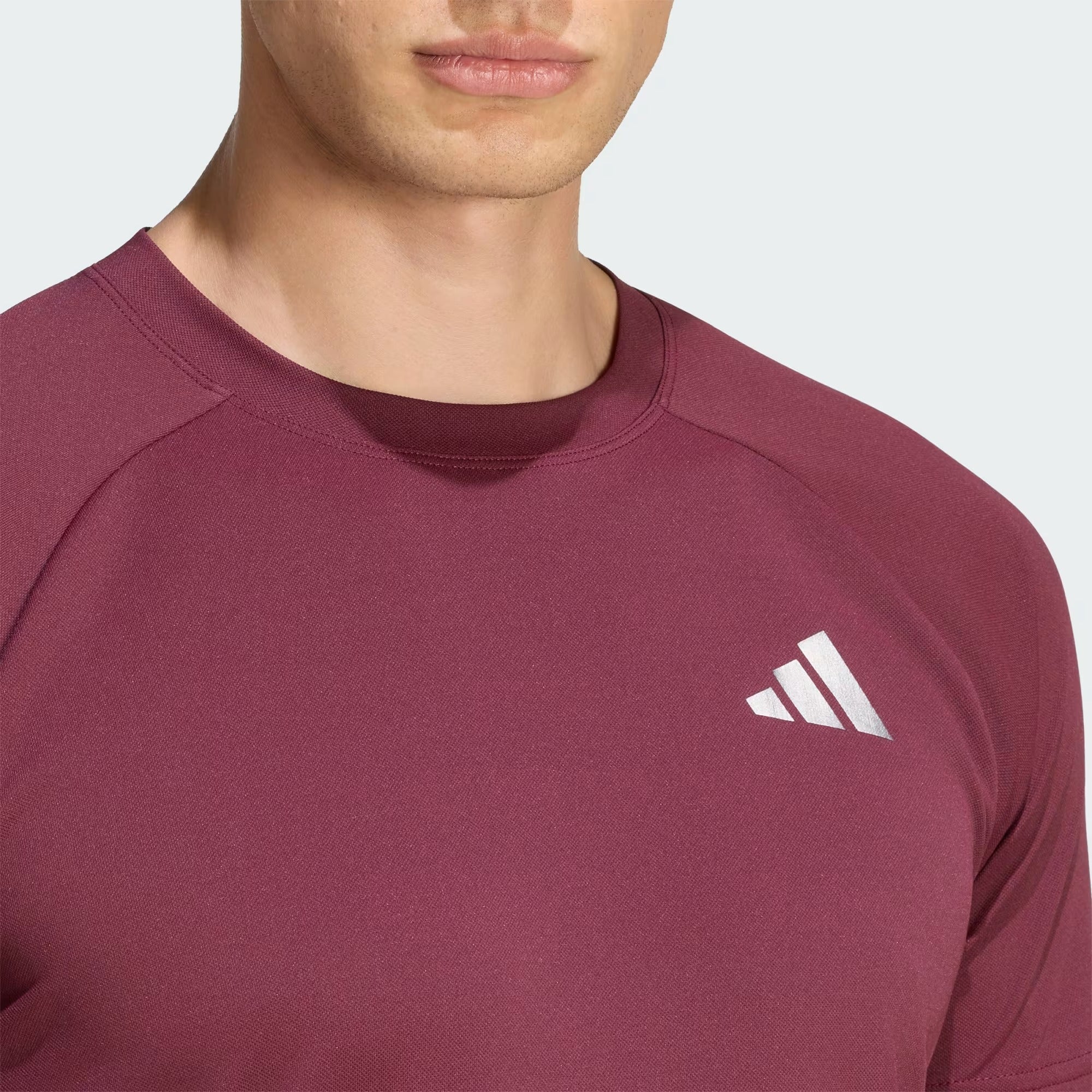 A person wearing the adidas Climacool+ Freelift Pro Men’s Badminton T-Shirt in maroon, featuring a white adidas logo on the chest, is shown from the shoulders up against a plain background.