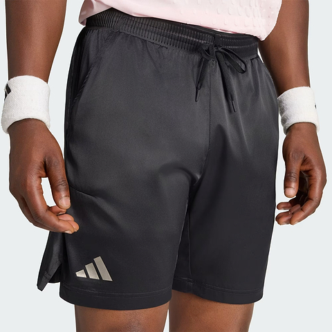 A person is wearing black adidas Climacool Ergo Pro Men’s Badminton Shorts, featuring an ergonomic cut, along with a light pink shirt and white wristbands. Only their lower torso and upper legs are visible as they stand with their hands by their sides.