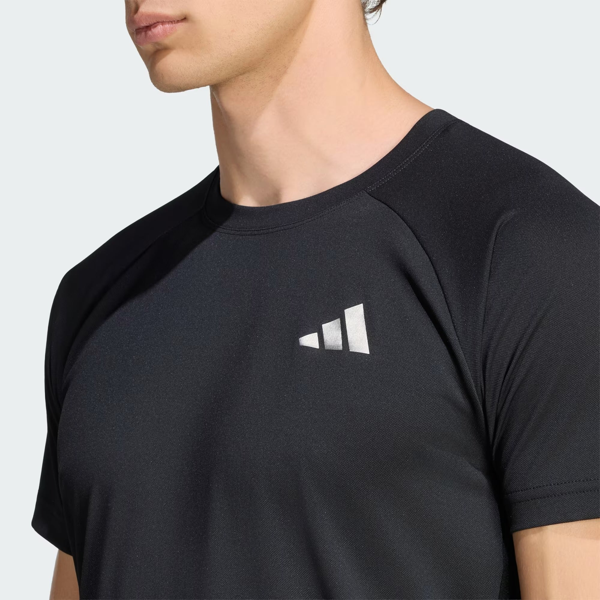 A person wearing the adidas Climacool+ Freelift Pro Men’s Badminton T-Shirt in black, featuring a white geometric logo on the left chest, is shown from the shoulders up against a plain background. This adidas performance badminton shirt incorporates advanced Climacool+ technology to provide superior cooling and comfort during intense matches.