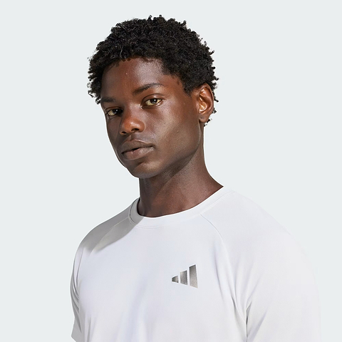A person with short curly hair is wearing the adidas ADIDAS Climacool+ Freelift Pro Men’s Badminton T-Shirt in white, which features a small silver geometric logo on the chest, while standing against a plain light background.