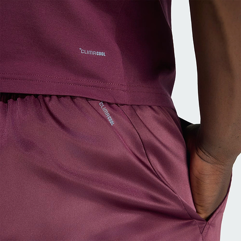 A close-up shows a person wearing maroon ADIDAS Climacool Ergo Pro Men’s Badminton Shorts, with their hand in the pocket. The word “Climacool” is visible on both the shirt and these ergonomic adidas badminton shorts. The background is plain and light-colored.