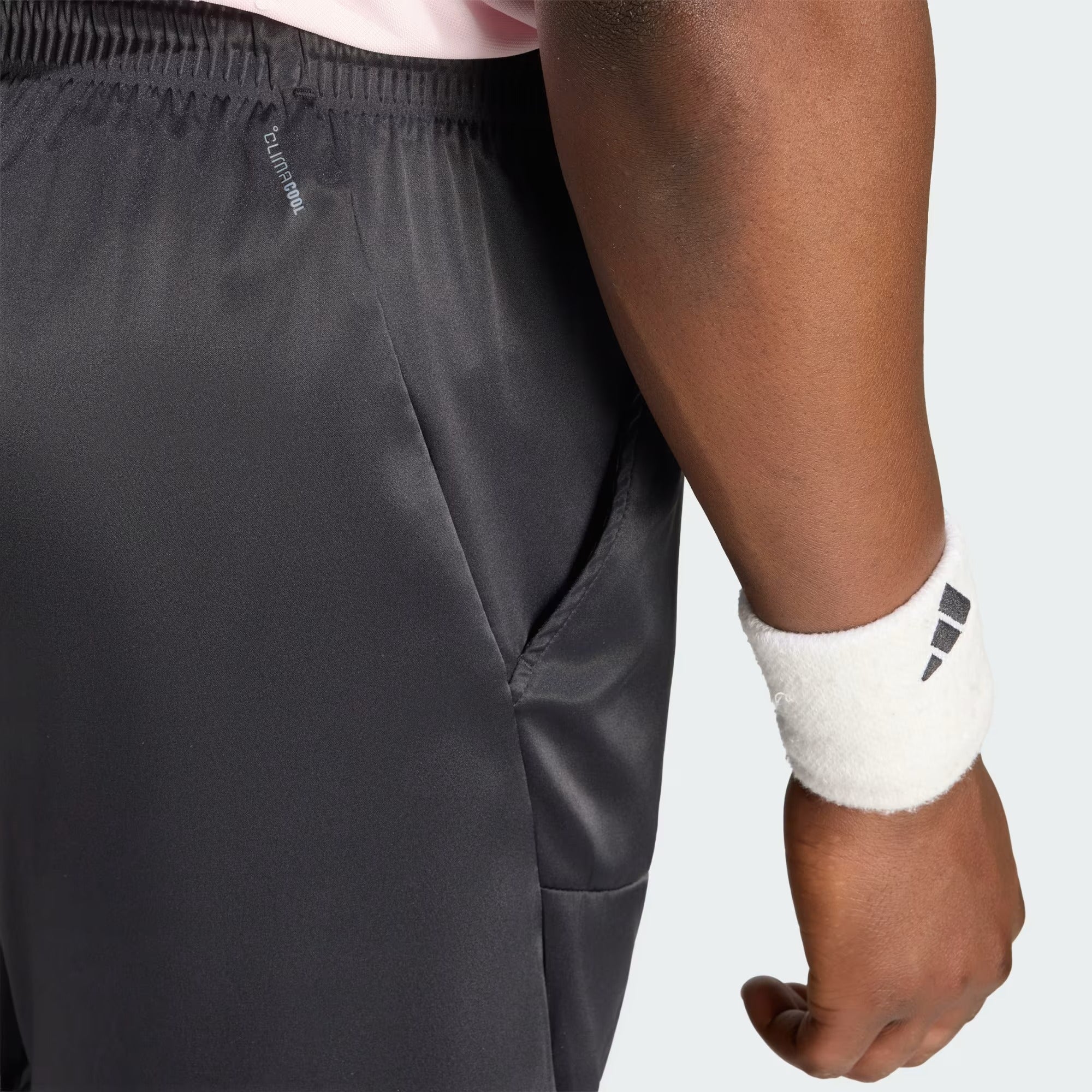 Close-up of a person wearing black ADIDAS Climacool Ergo Pro Men’s Badminton Shorts featuring ergonomic construction and a visible pocket, paired with a white adidas wristband and a pink shirt partially visible at the waist.