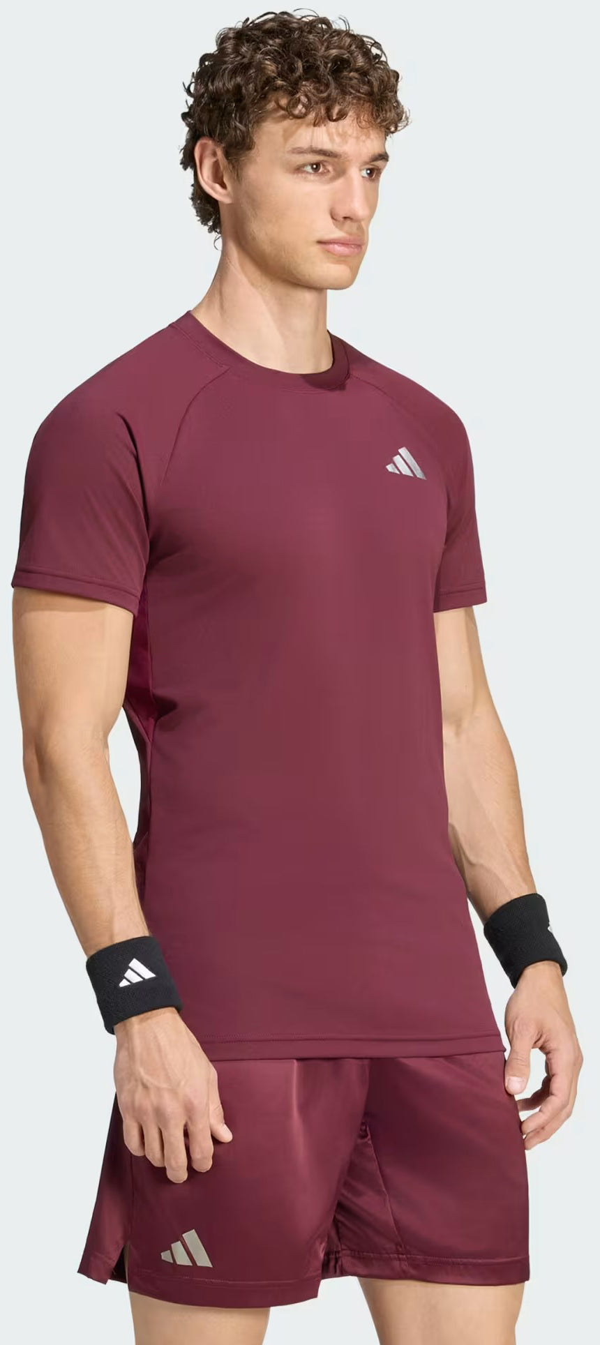 A man stands facing forward, dressed in the adidas Climacool+ Freelift Pro Men’s Badminton T-Shirt in maroon and matching shorts, both featuring adidas logos. He wears black adidas wristbands on each wrist, highlighting his sporty style in this performance tee enhanced with ADIDAS Climacool+ technology.