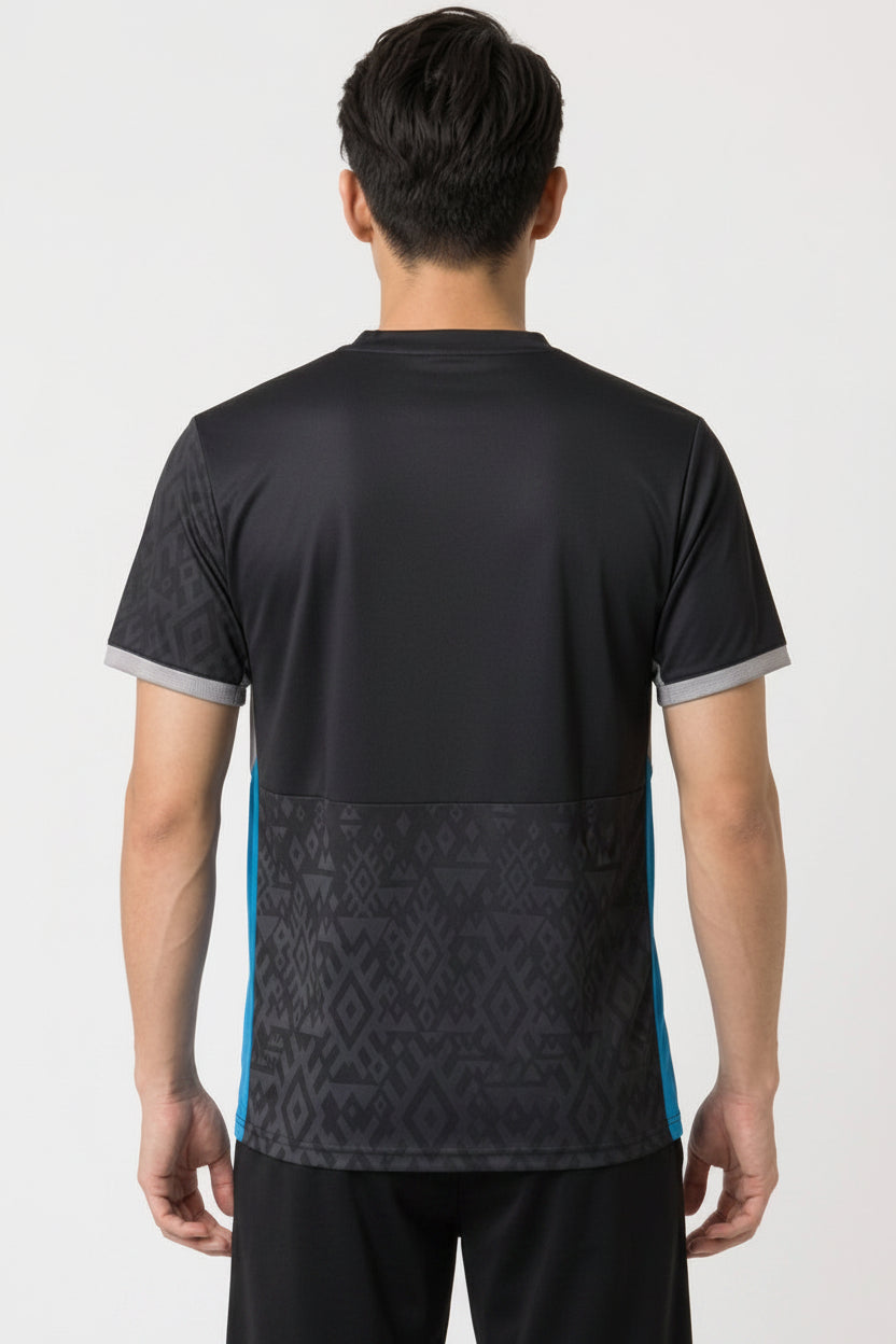 A man with short black hair stands with his back to the camera, wearing the Yonex YTM11 Mens Badminton T-Shirt in black, featuring geometric patterns. This Yonex shirt offers quick-dry comfort for intense matches.