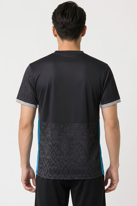 A man with short black hair stands with his back to the camera, wearing the Yonex YTM11 Mens Badminton T-Shirt in black, featuring geometric patterns. This Yonex shirt offers quick-dry comfort for intense matches.