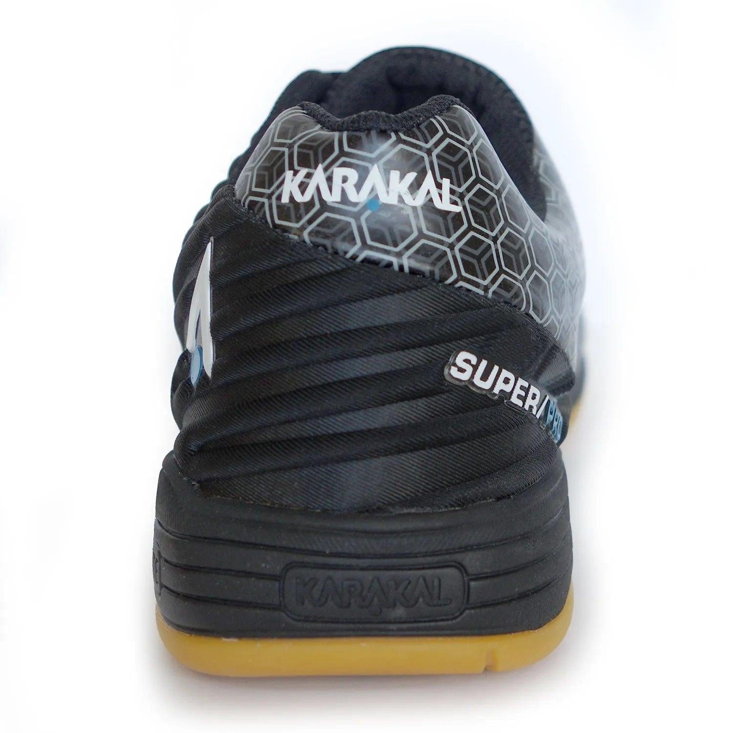 The image presents the rear view of the black Karakal Super Pro Indoor Court Badminton Shoes, highlighting an asymmetric lace system and hexagonal design, accompanied by "SUPER" text on the side. These indoor court shoes combine style and performance with a gum-colored sole and prominently feature the Karakal brand name on both the heel and sole.