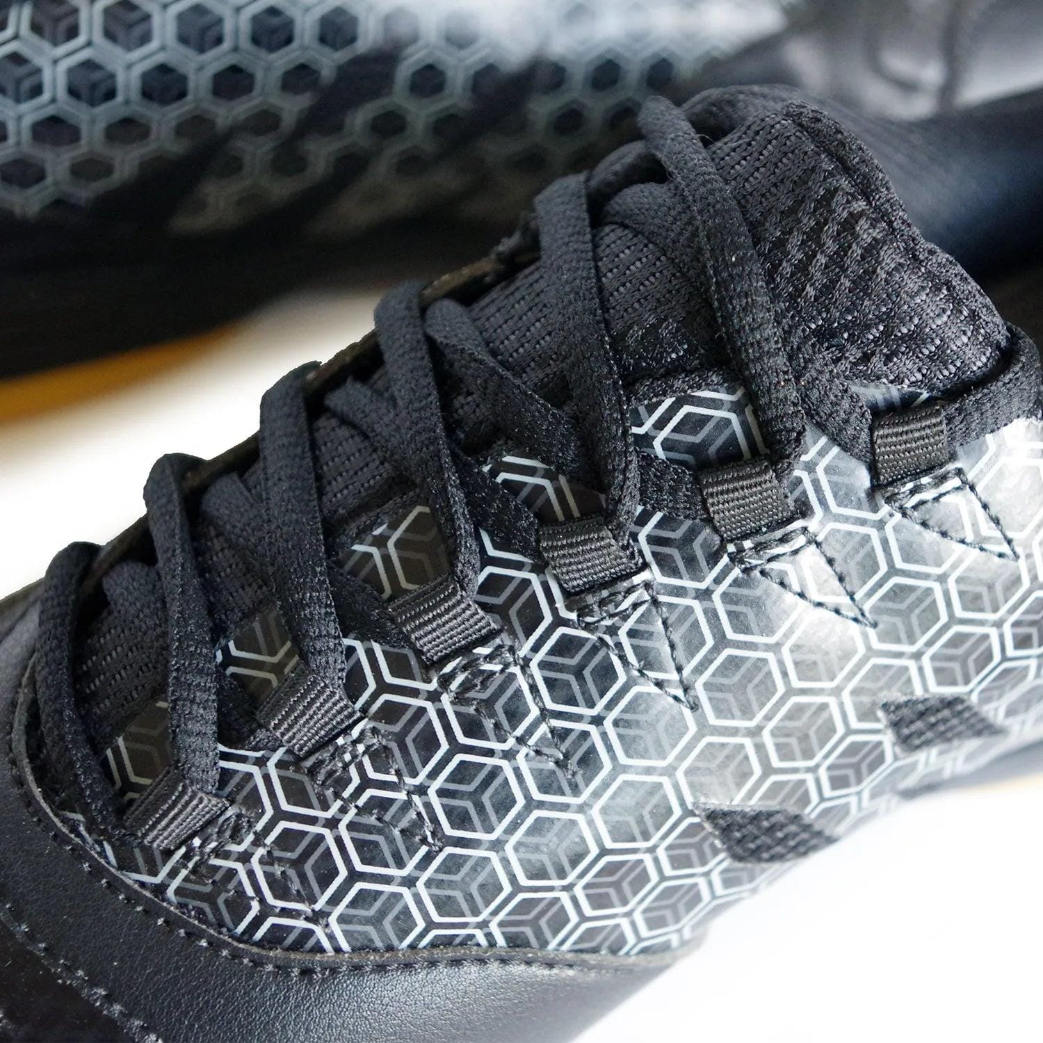 A close-up of the Karakal Super Pro Indoor Court Badminton Shoes showcases a sleek black design with a prominent white geometric hexagonal pattern. The asymmetric lace system, complemented by black laces, emphasizes the detailed upper part, reminiscent of high-performance indoor court footwear.