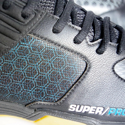 A close-up of the Karakal Super Pro Indoor Court Badminton Shoes in black showcases a striking blue hexagonal pattern on the side panel, with "SUPER/PRO" emblazoned in white. These indoor court shoes feature an asymmetric lace system with black laces, complemented by a ribbed heel design and a light-colored sole at the bottom.